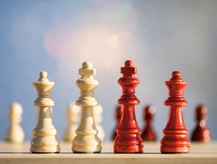 A stock photo of four chess pieces lined up with several pawns lined up in the background. The two chess pieces on the left are white. and the two chess pieces on the right are red.