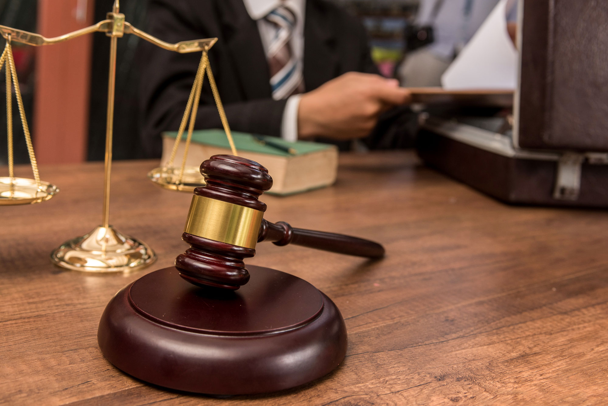 A stock photo of a table with a gavel, the scales of justice, a book, and an open briefcase that a lawyer is taking a file out of