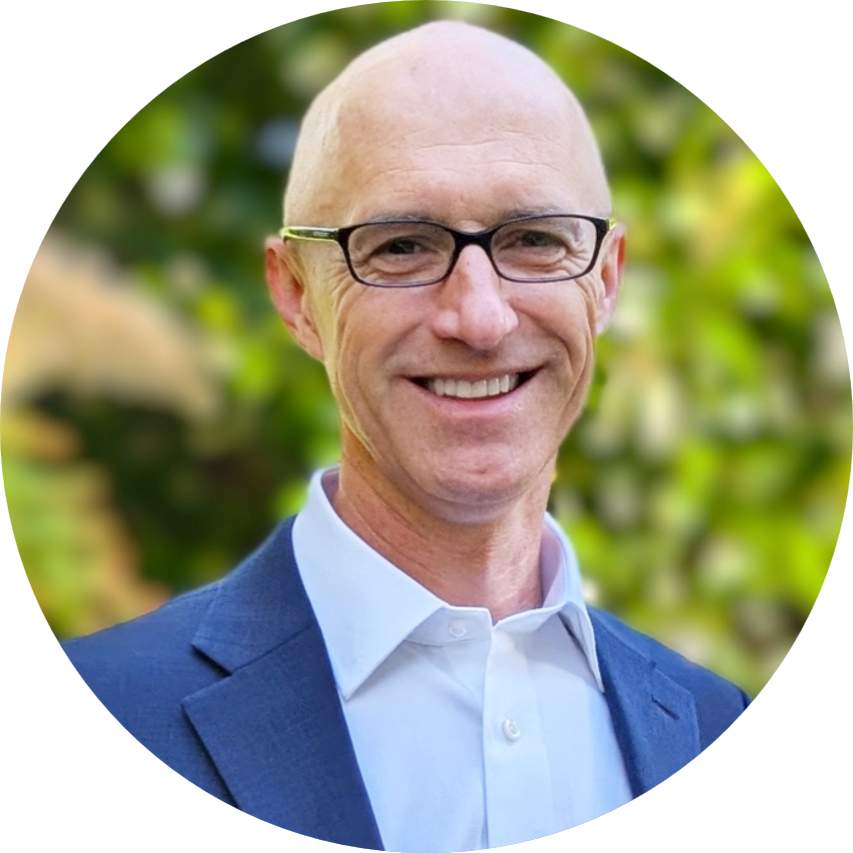 Smiling bald man in glasses, white shirt, and blue suit jacket against blurred green background.